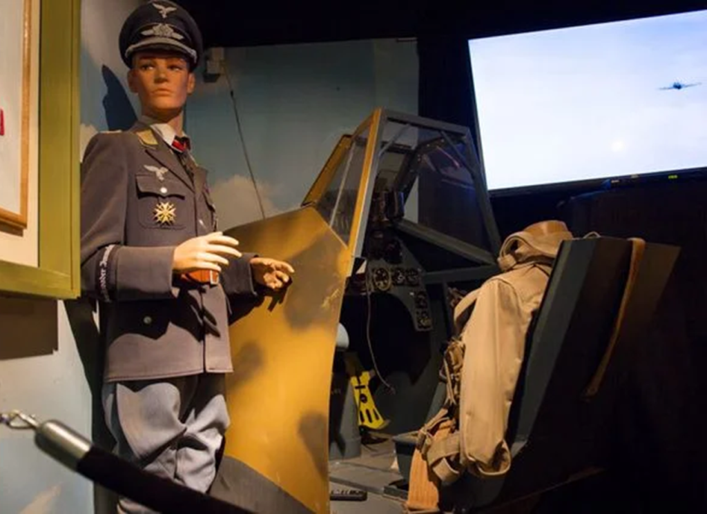 Outdoor daytime photo of a full-scale Harrier Jump Jet display at the Volo Museum, showing the military aircraft with its angled wings and vertical takeoff capability, positioned on a paved exhibit area with visitors in the background