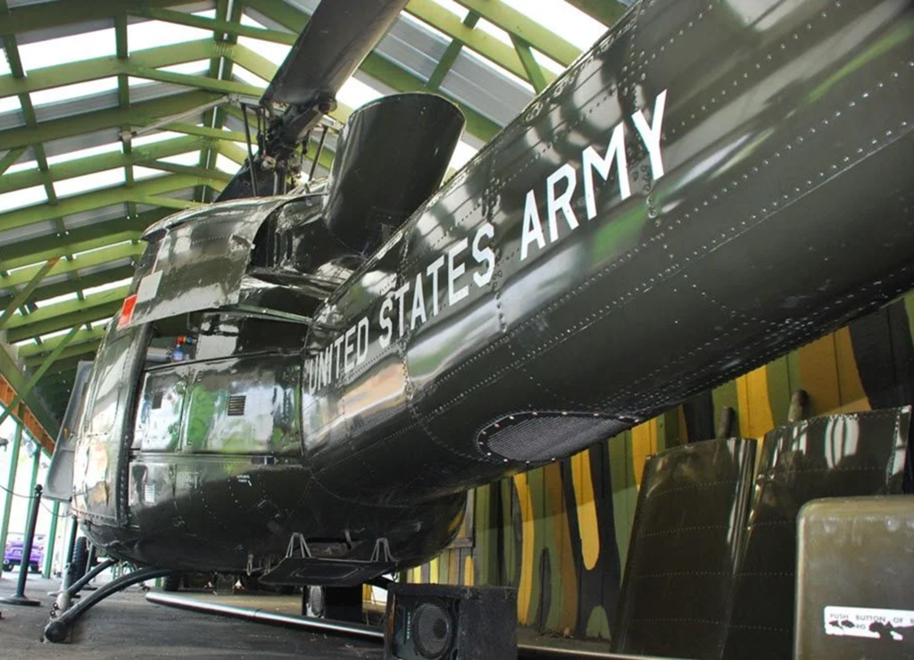 Museum exhibit showing a dark green United States Army helicopter with ‘UNITED STATES ARMY’ text on the fuselage, parked indoors beneath a metal roof structure with visible rotor blades above.