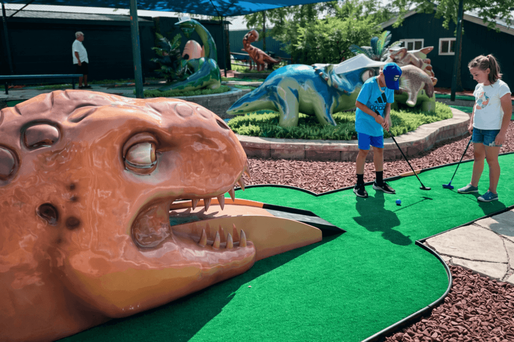Kids playing mini-golf with dinosaur sculptures.